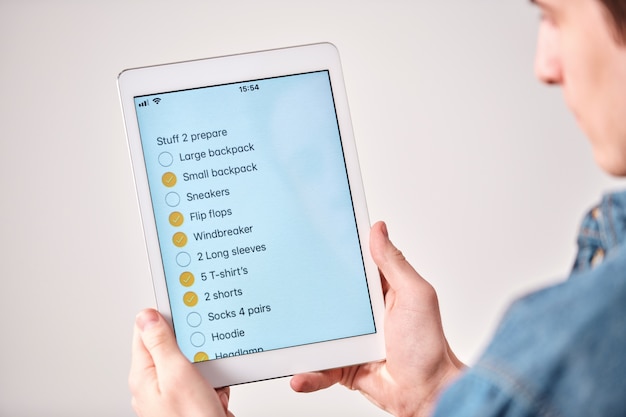 Photo sur l'épaule du jeune homme à l'aide de tablette et liste de contrôle lors de l'emballage de la valise pour le voyage