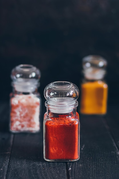 Photo Épices aromatiques lumineuses dans des bocaux en verre, assortiments d'épices : sel rose, paprika et curcuma sur fond de pierre noire.