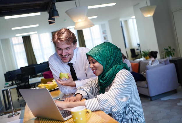 équipe d'affaires multiculturelle internationale.homme mangeant une pomme femme musulmane africaine portant le hijab vert buvant du thé tout en travaillant ensemble sur un ordinateur portable dans un espace de détente au démarrage moderne à plan ouvert