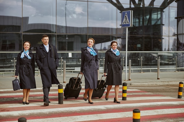 L'équipe d'avion marche avec des bagages sur un passage pour piétons près de l'aéroport moderne. Les jeunes femmes souriantes et l'homme portent l'uniforme. Hôtesse de l'air et hôtesses de l'air. Travail en équipe. Aviation civile commerciale. Concept de voyage aérien