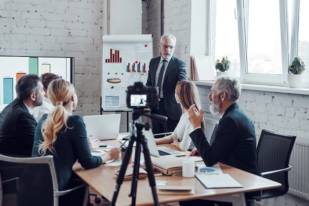 Une équipe commerciale prospère écoute un homme qui fait une présentation tout en filmant une réunion du personnel dans la salle du conseil