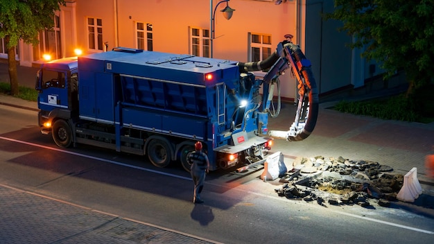 Une équipe de constructeurs de routes effectue des réparations routières la nuit à l'aide d'un gros aspirateur de camion Des réparateurs professionnels changent le trou d'homme sur la chaussée