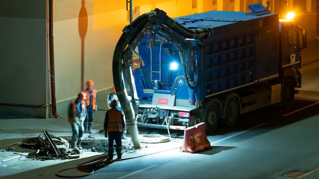 Une équipe de constructeurs de routes effectue des réparations routières la nuit à l'aide d'un gros aspirateur de camion Des réparateurs professionnels changent le trou d'homme sur la chaussée