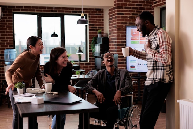 Photo une équipe diversifiée de travailleurs souriants et d'hommes paralysés prenant une pause du travail dans un bureau d'affaires. employé de sexe masculin en fauteuil roulant souffrant de problèmes de santé physique riant avec des collègues.