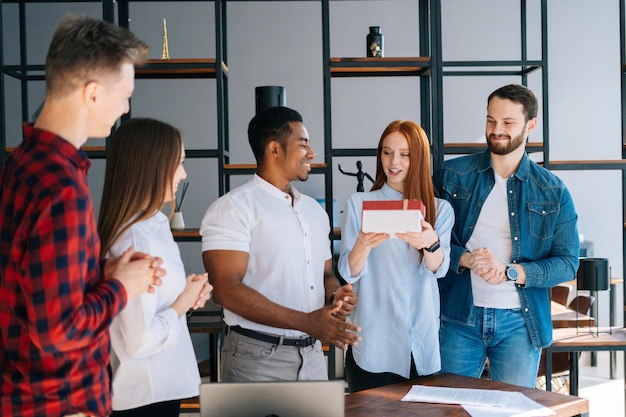 Une équipe heureuse d'hommes d'affaires multiethniques félicite un collègue afro-américain et offre un cadeau avec surprise. Un joyeux anniversaire est surpris d'ouvrir la boîte avec un cadeau sous les applaudissements de ses collègues.