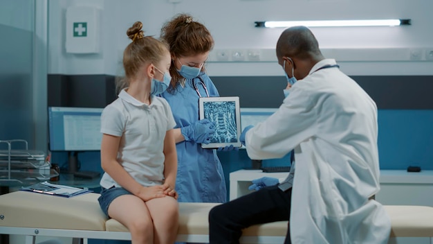 Une équipe médicale diversifiée expliquant le diagnostic de l'analyse par rayons X à la petite fille lors du contrôle pendant la pandémie de covid 19. Infirmière et médecin montrant les résultats de l'examen de radiographie à l'enfant lors d'un rendez-vous de consultation.