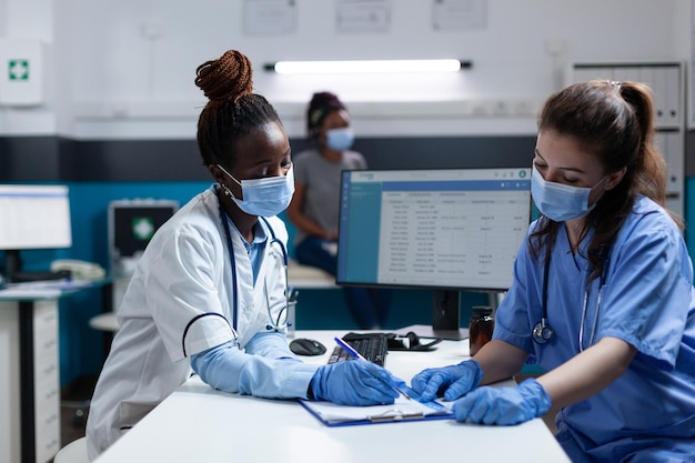 Équipe médicale avec masque protecteur pour prévenir l'infection par covid19 écrivant une prescription de médicaments lors d'un rendez-vous clinique au bureau de l'hôpital. Médecin afro-américain examinant un patient malade