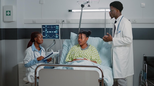Équipe médicale parlant à une femme malade dans un lit d'hôpital. Homme et femme travaillant comme médecins faisant des consultations de soins de santé. Jeune patient avec poche intraveineuse et moniteur de fréquence cardiaque en convalescence