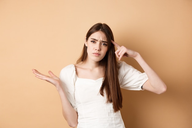 Es-tu stupide. Femme agacée se moquant de la personne, pointant du doigt la tête et haussant les épaules confuse, irritée par des idées folles, debout sur fond beige.