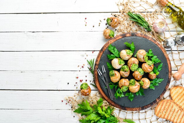 Escargots cuits au four avec du beurre à l'ail et des herbes fraîches sur une plaque noire sur un fond en bois blanc Vue de dessus Espace libre pour votre texte