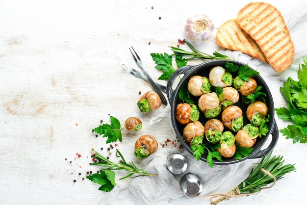 Escargots cuits au four avec du beurre à l'ail et des herbes fraîches sur une plaque noire sur un fond en bois blanc Vue de dessus Espace libre pour votre texte