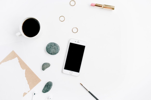 Espace de travail de bureau à domicile avec café, téléphone portable, enveloppe artisanale, pierres, écouteurs et papier vierge sur une surface blanche