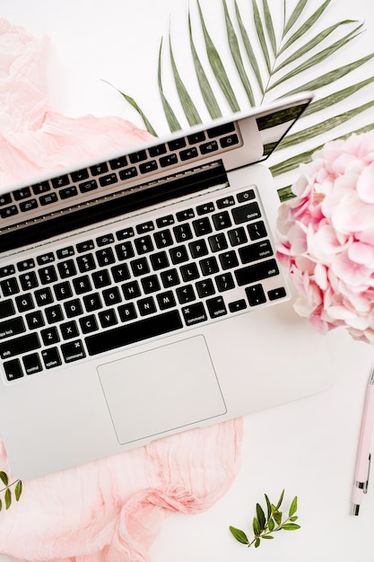 Espace de travail de bureau à domicile femme avec ordinateur portable, bouquet de fleurs d'hortensia rose, couverture pastel, plaque de feuille de monstera et accessoires sur surface blanche