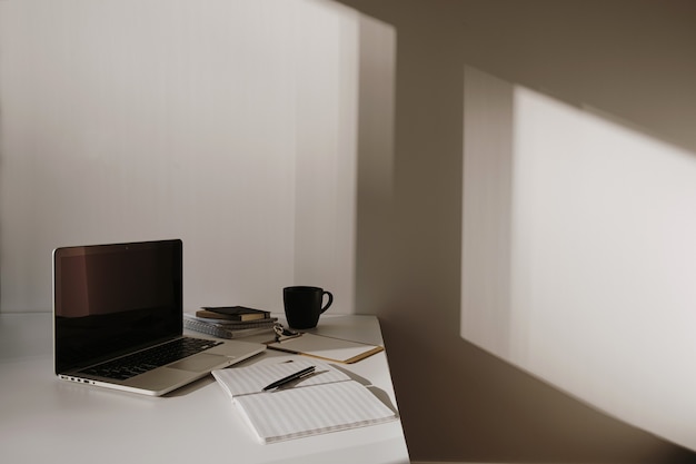 Espace de travail de bureau à domicile minimaliste. Ordinateur portable sur table avec tasse à café, feuille de papier, papeterie contre mur blanc