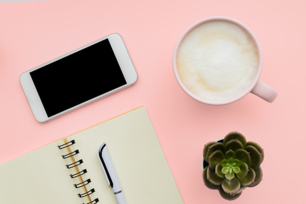 Espace de travail de bureau Feminini avec smartphone, bloc-notes vierge, stylo, tasse de café sur le mur rose. mise à plat, vue de dessus