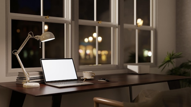 Un espace de travail de bureau moderne la nuit avec une maquette d'ordinateur portable à écran blanc sur une table contre la fenêtre