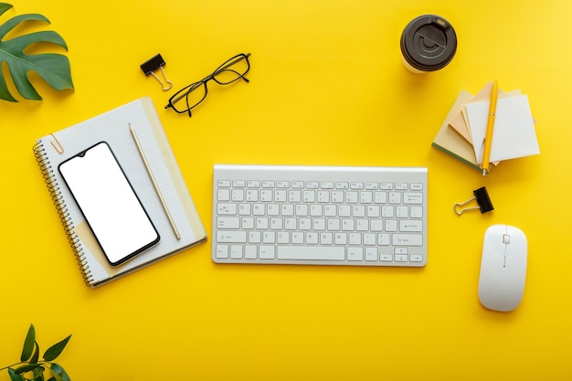 Espace de travail de bureau plat sur fond jaune coloré. Table de bureau avec clavier d'ordinateur, lunettes, souris pour smartphone, plantes de fournitures de bureau, tasse à café. Écran de téléphone maquette à plat.