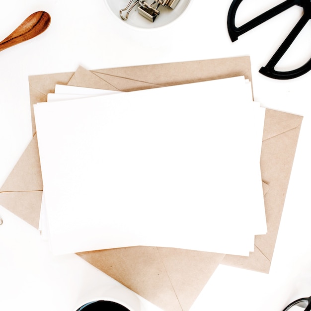 Espace de travail avec du papier vierge, du café, une enveloppe artisanale, des ciseaux, des fournitures de bureau sur une surface blanche