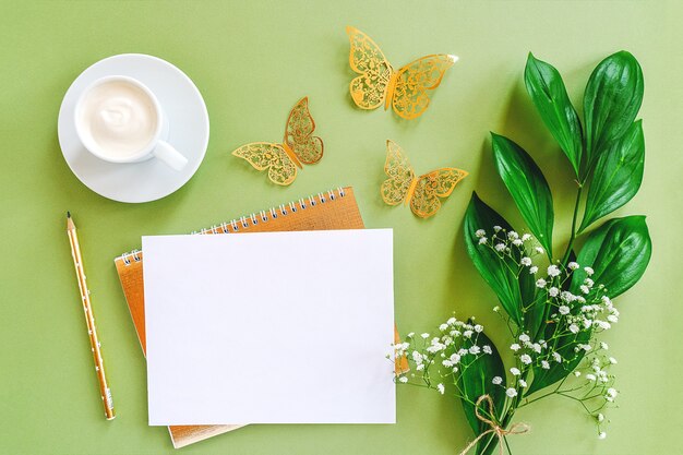 Espace de travail avec une feuille vierge de bloc-notes, un crayon, une feuille verte, des papillons dorés décoratifs et une tasse de café sur fond vert