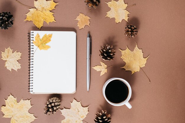 Espace de travail avec des feuilles d'érable dorées, tasse à café, bosses, cahier et stylo sur fond marron.