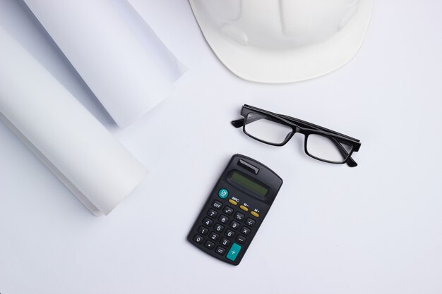 Espace de travail de l'ingénieur. Casque de construction, rouleaux de dessins, calculatrice et lunettes sur fond blanc. Vue de dessus. Copier l'espace