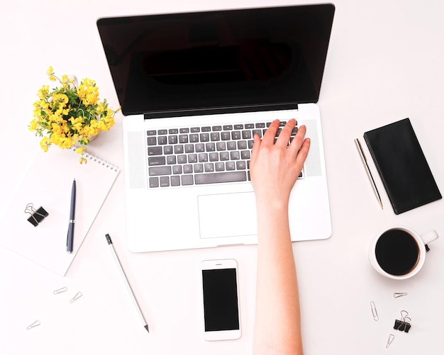 Espace de travail avec main de femme sur clavier d'ordinateur portable, téléphone portable, café, verres et fleurs sur fond blanc. Mise à plat, vue de dessus