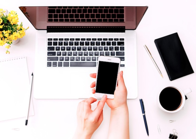 Espace De Travail Avec Une Main De Femme Tenant Un Téléphone Portable Sur Un Clavier D'ordinateur Portable, Du Café Et Des Fleurs Jaunes Sur Fond Blanc. Mise à Plat, Vue De Dessus