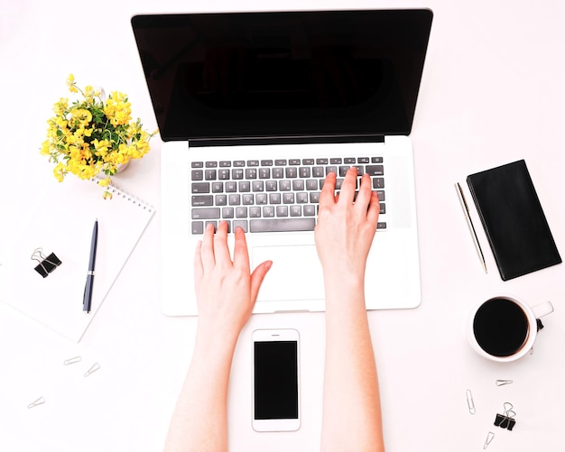 Espace de travail avec des mains de femme sur un clavier d'ordinateur portable, un téléphone portable, du café, des verres et des fleurs sur fond blanc. Mise à plat, vue de dessus