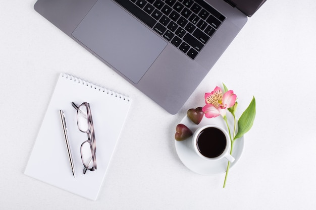 Espace de travail avec ordinateur portable, bloc-notes, tablette, verres, tasse de café noir et une fleur de lis péruvienne sur fond blanc