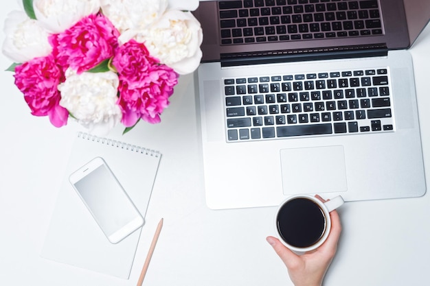 Espace de travail avec ordinateur portable, téléphone portable, tasse de café et main féminine, carnet, stylos et fleurs de pivoine roses et blanches sur fond blanc. Mise à plat, vue de dessus, lieu de travail indépendant