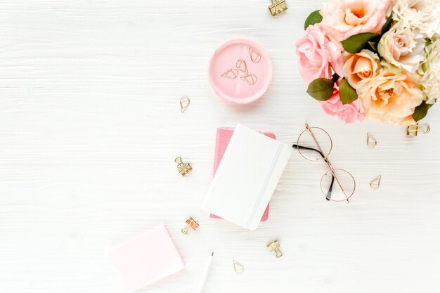 Espace de travail pour femmes avec des accessoires de bouquet de fleurs de roses roses et beiges lunettes de journal sur blanc