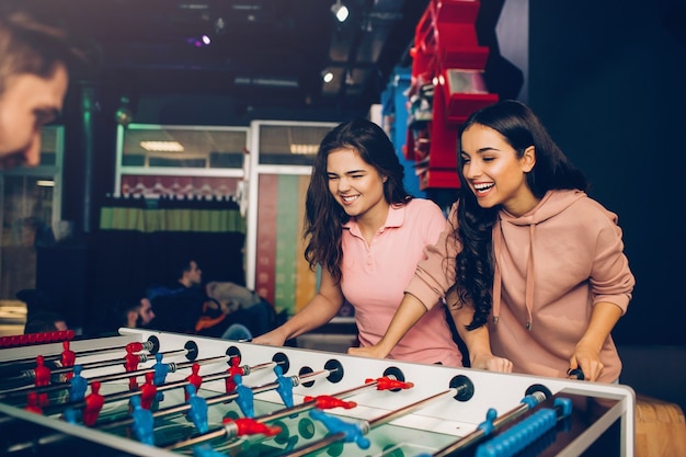 Espiègles jeunes femmes excitées jouant au baby-foot avec guy dans la chambre. Les mannequins jouent au jeu et le contrôlent.