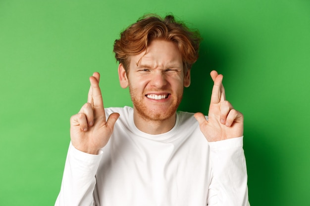 Espoir homme de race blanche à la recherche de soucis, en attente de résultats importants et en faisant le vœu avec les doigts croisés, debout sur fond vert.