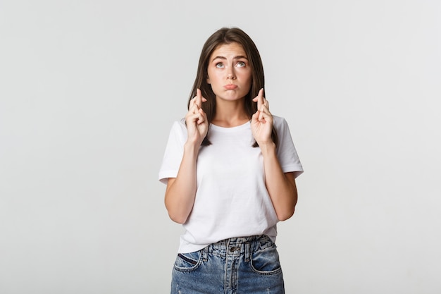 Espoir jolie fille brune croise les doigts bonne chance, levant les yeux et faisant un vœu sur blanc.