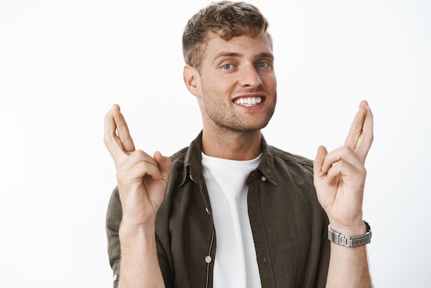 Espoir et optimiste bel homme aux cheveux blonds croise les doigts pour la bonne chance priant et faisant le vœu de rêver devenu réalité souriant ravi anticipant avec foi sur fond gris