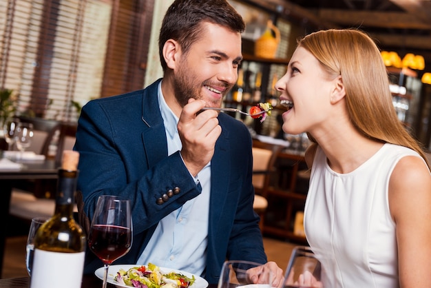 Essaye ça! Beau jeune couple d'amoureux en train de dîner au restaurant pendant que l'homme nourrit sa petite amie avec de la salade