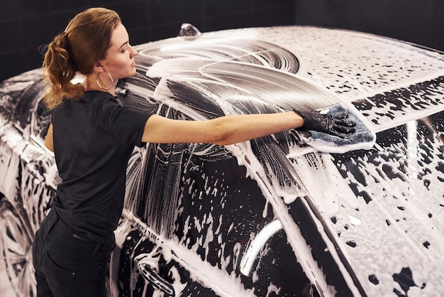 Essuie le véhicule qui est dans du savon blanc L'automobile noire moderne est nettoyée par une femme à l'intérieur de la station de lavage de voiture