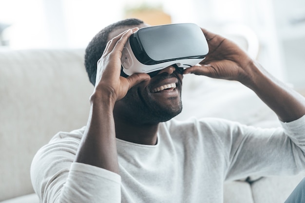 C'est cool! Beau jeune homme africain ajustant son casque VR et souriant assis sur le tapis à la maison