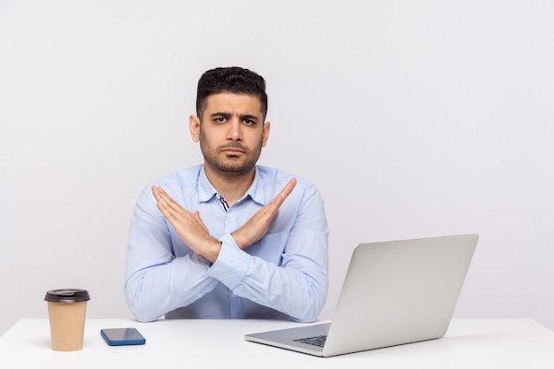 C'est la finition de fin Patron strict assis au bureau avec un ordinateur portable sur le bureau croisant les mains montrant le signe x geste d'arrêt avertissant des problèmes tourné en studio à l'intérieur isolé sur fond blanc