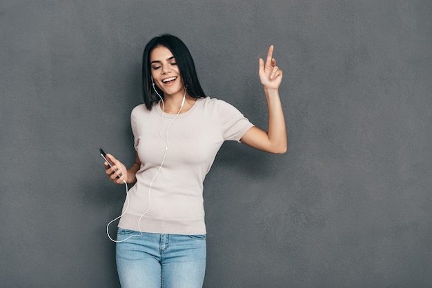 C'est l'heure de la musique ! Jolie jeune femme souriante dans les écouteurs tenant un téléphone intelligent et faisant des gestes en se tenant debout sur fond gris