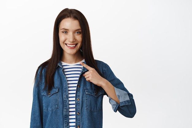 C'est moi. Jeune femme souriante, se montrant du doigt, nommant, se portant volontaire pour faire qch, se vanter ou se promouvoir, parlant de réalisation personnelle, debout sur fond blanc.