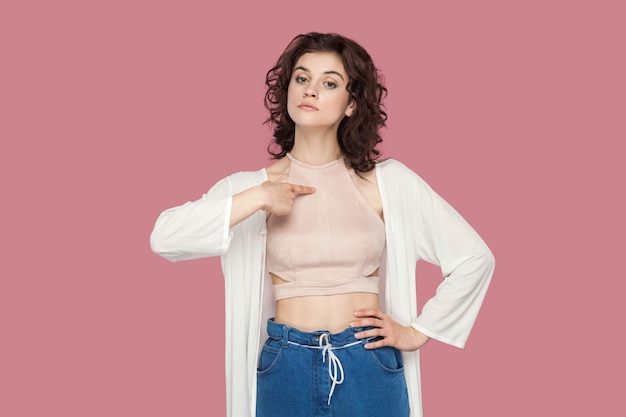 C'est moi. Portrait d'une belle jeune femme brune fière et hautaine avec une coiffure frisée dans un style décontracté debout, se pointant et regardant la caméra. tourné en studio isolé sur fond rose.