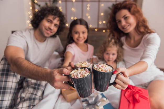 C'est Noël! Des parents heureux et leurs filles tiennent des tasses et sourient assis sur le lit avec des cadeaux