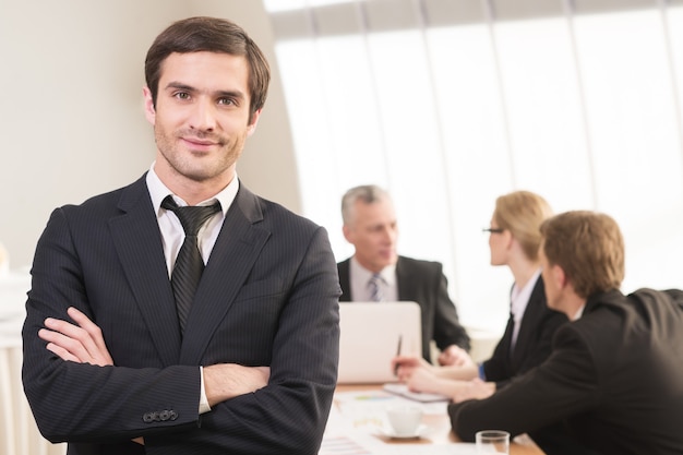 C'est un vrai leader. Beau jeune homme en tenue de soirée gardant les bras croisés et souriant pendant que ses collègues travaillent en arrière-plan