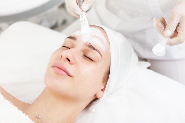 Esthéticienne applique un masque sur un beau visage féminin dans un salon de beauté Hygiène du visage procédures cosmétiques soins de la peau nettoyage des pores de la peau concept de cosmétologie