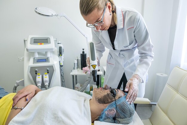 L'esthéticienne ou le dermatologue applique un masque blanc sur le visage du client avec une brosse