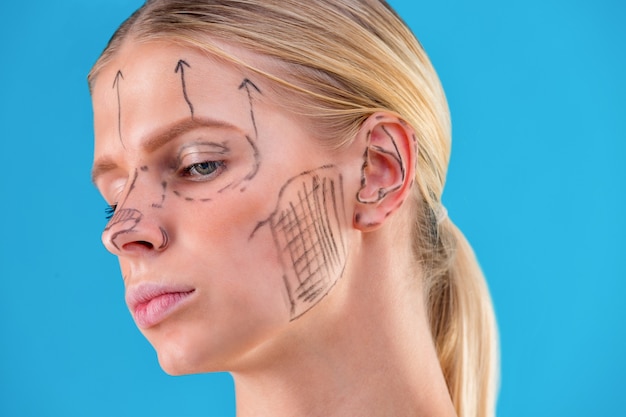 Esthéticienne dessiner des lignes de correction sur le visage de la femme. Avant l'opération de chirurgie plastique. Isolé sur bleu.