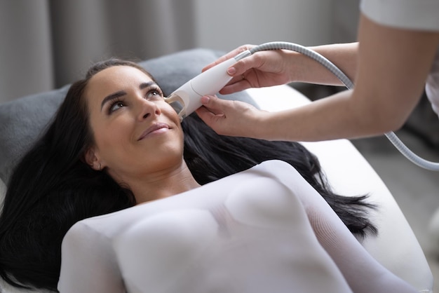 Photo l'esthéticienne fournit des soins du visage à une belle femme allongée dans le studio de cosmétiques par un appareil électronique