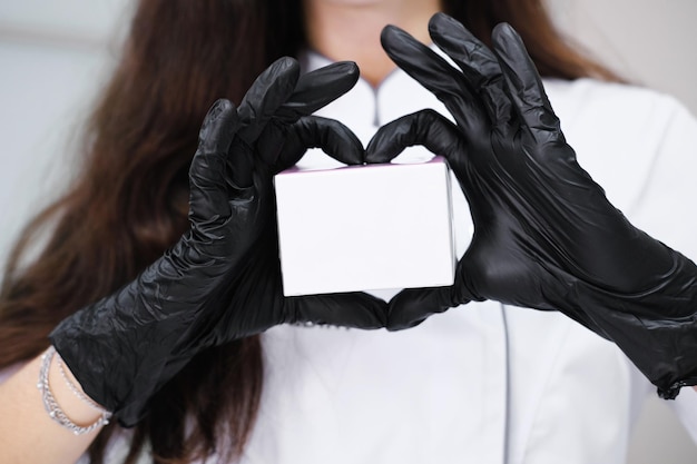 L'esthéticienne en gants noirs fait un signe de coeur autour de la crème dans l'emballage Concept de soins de la peau