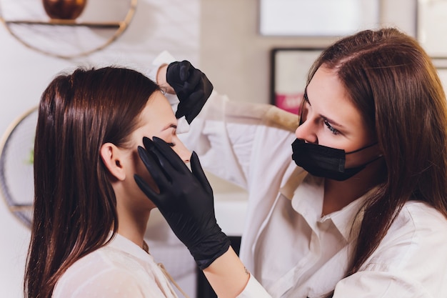 Esthéticienne-maquilleuse applique de la peinture au henné sur des sourcils préalablement épilés, dessinés et taillés dans un salon de beauté lors de la correction de la séance. Soin professionnel du visage.